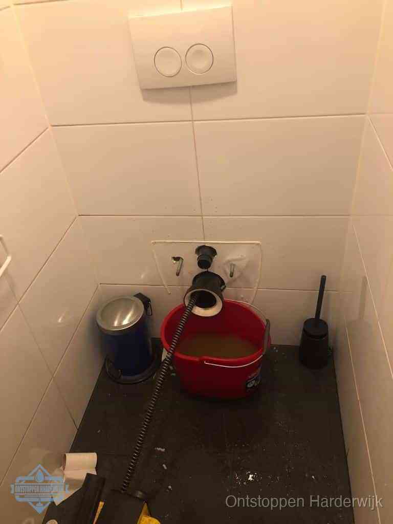 Een ontstopte wand-wc met verwijderde toiletpot waar een ontstoppingsveer in de wanduitgang verdwijnt, het achterhalen waar de verstopping zit in Harderwijk