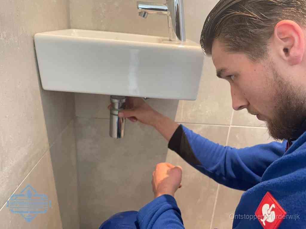 Een monteur draait met de hand de zwanenhalssifon onder een fonteinwasbak in de toiletruimte los om de bron van rioollucht in Harderwijk te vinden