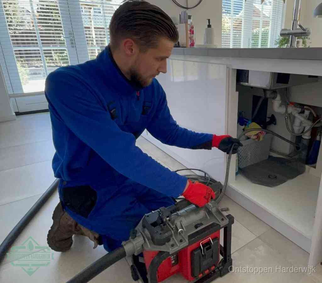 Een monteur met rode handschoenen knielt in een keuken en voert een elektrische ontstoppingsveer in tijdens het professioneel afvoer ontstoppen in Harderwijk